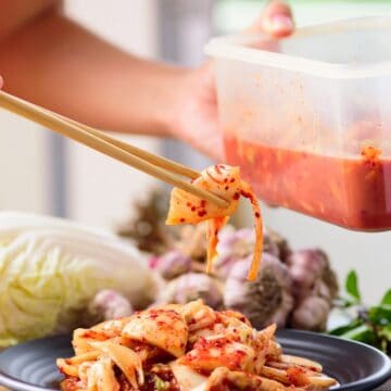 A person uses chopsticks to serve kimchi from a plastic container onto a black plate. A head of cabbage, garlic bulbs, and leafy herbs are visible on the table. The background is softly blurred.
