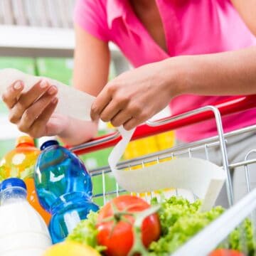 Person in a pink shirt checks a long receipt while standing next to a shopping cart filled with groceries like bottled drinks, lettuce, celery, and tomatoes, in a store aisle. Shelves stocked with products are visible in the background.