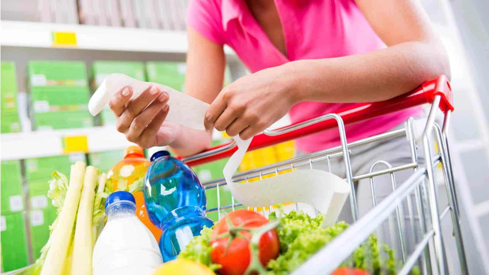 Person in a pink shirt checks a long receipt while standing next to a shopping cart filled with groceries like bottled drinks, lettuce, celery, and tomatoes, in a store aisle. Shelves stocked with products are visible in the background.