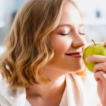 A person with shoulder-length hair is smiling with eyes closed, holding a green apple close to their face. They appear to be enjoying the apple's aroma. The background is a softly focused kitchen setting.