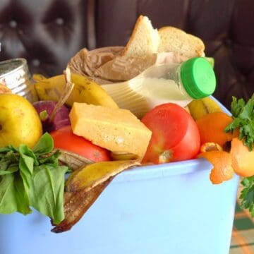 A blue container filled with mixed food items, including leafy greens, apples, bananas, oranges, a can, a loaf of bread, a paper bag, a bottle, and cheese. The container is on a checkered tablecloth with a padded background.