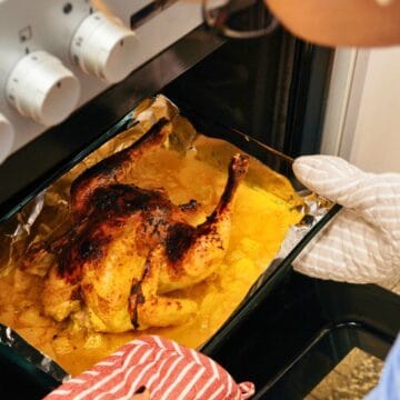 A person is placing a roasted chicken into an oven on a foil-lined baking sheet. They are wearing a quilted oven mitt and holding a red-striped towel. The oven has three white dials on the side. The chicken appears to be cooked with a golden-brown surface.