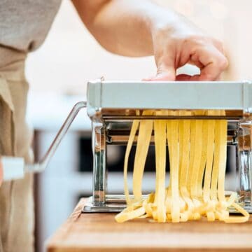 A person uses a hand-cranked pasta machine to cut fresh pasta dough into noodles. The machine is on a wooden counter. There is a bottle of olive oil and an egg nearby. The background is softly blurred.