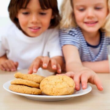 Two children reach for cookies on a plate. They are sitting at a table with glasses of milk. The child on the left has brown hair and is wearing a white shirt; the child on the right has blonde hair and is wearing a striped shirt.