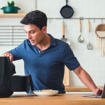 A man in a blue shirt is using an air fryer on a kitchen counter. A laptop is open next to him, and a plate with food is in front of him. Kitchen utensils hang on the wall behind.