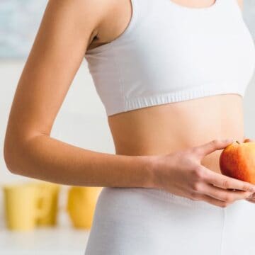 A person in a white sports outfit holds a peach in a kitchen. The background features wooden utensils and stacked plates. The setting suggests a modern kitchen environment.