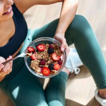 A woman in athletic wear sits on the floor holding a glass bowl with cereal and berries. Beside her, there are blue dumbbells. She is looking away and smiling. The scene suggests a post-workout snack.