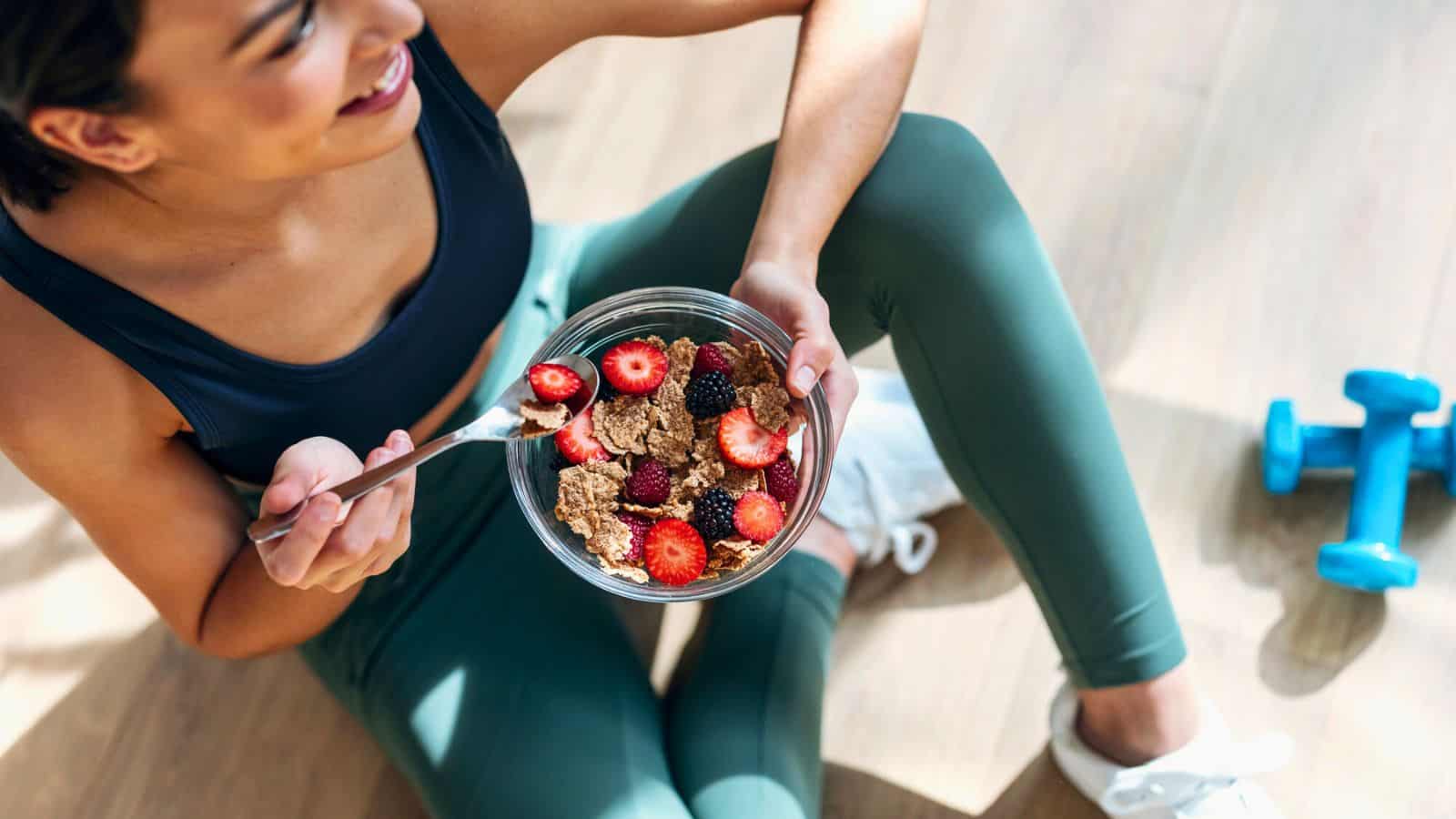 A woman in athletic wear sits on the floor holding a glass bowl with cereal and berries. Beside her, there are blue dumbbells. She is looking away and smiling. The scene suggests a post-workout snack.