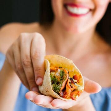 A woman holds a taco in her hand, with ingredients like meat and vegetables visible. She is smiling, wearing an off-the-shoulder blue top, and has dark hair. The background is dark and out of focus.