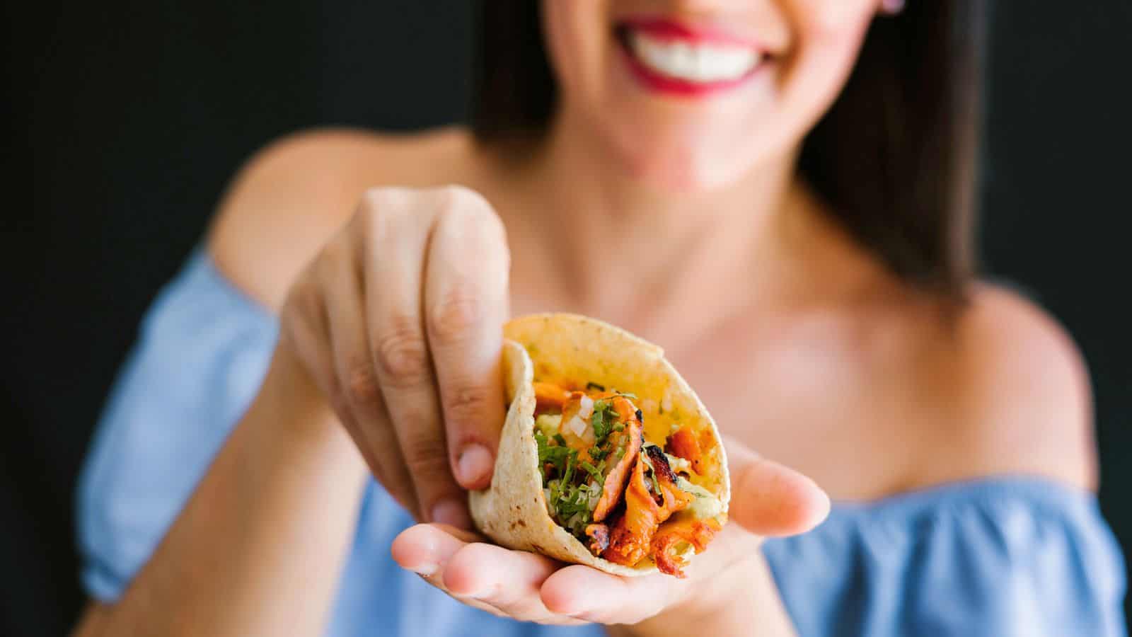 A woman holds a taco in her hand, with ingredients like meat and vegetables visible. She is smiling, wearing an off-the-shoulder blue top, and has dark hair. The background is dark and out of focus.