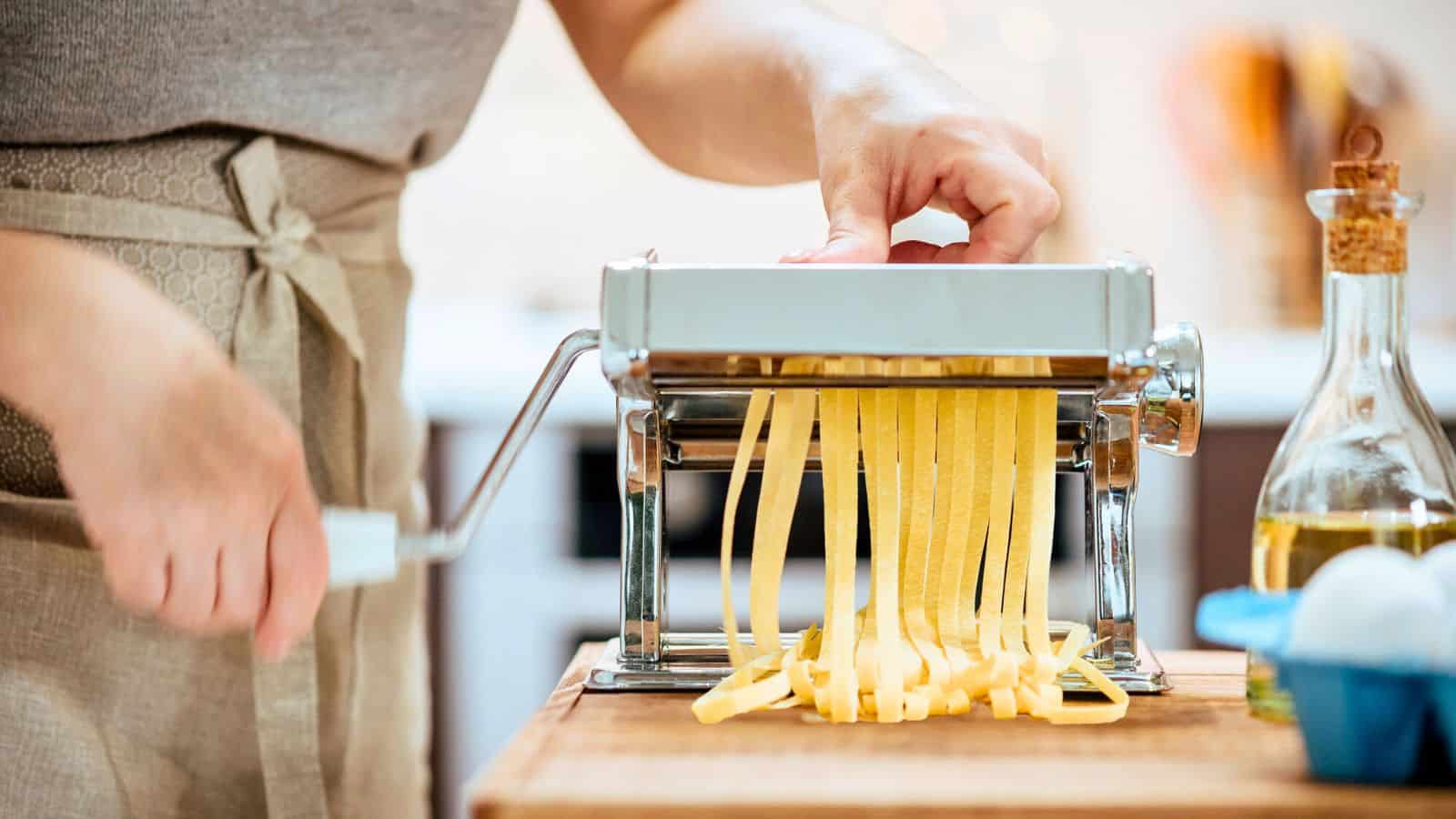 A person uses a hand-cranked pasta machine to cut fresh pasta dough into noodles. The machine is on a wooden counter. There is a bottle of olive oil and an egg nearby. The background is softly blurred.