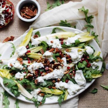 A salad on a white oval plate features endive, arugula, crumbled cheese, pomegranate seeds, and pecans. A half pomegranate, a small bowl of pecans, and a knife are nearby on a cloth-covered surface.