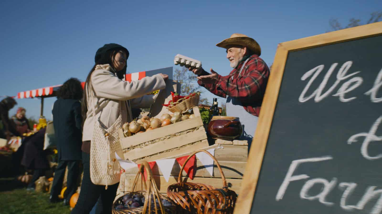 A marketplace scene shows a person handing a carton of eggs to another person across a wooden display of vegetables. A chalkboard with writing is partially visible on the right. Other people and market stalls are in the background under a clear sky.
