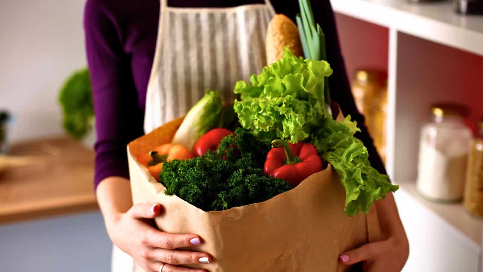 A person holds a paper bag filled with fresh vegetables, including lettuce, red bell pepper, kale, zucchini, and a baguette. The person wears a striped apron, and kitchen shelves with jars are visible in the background.