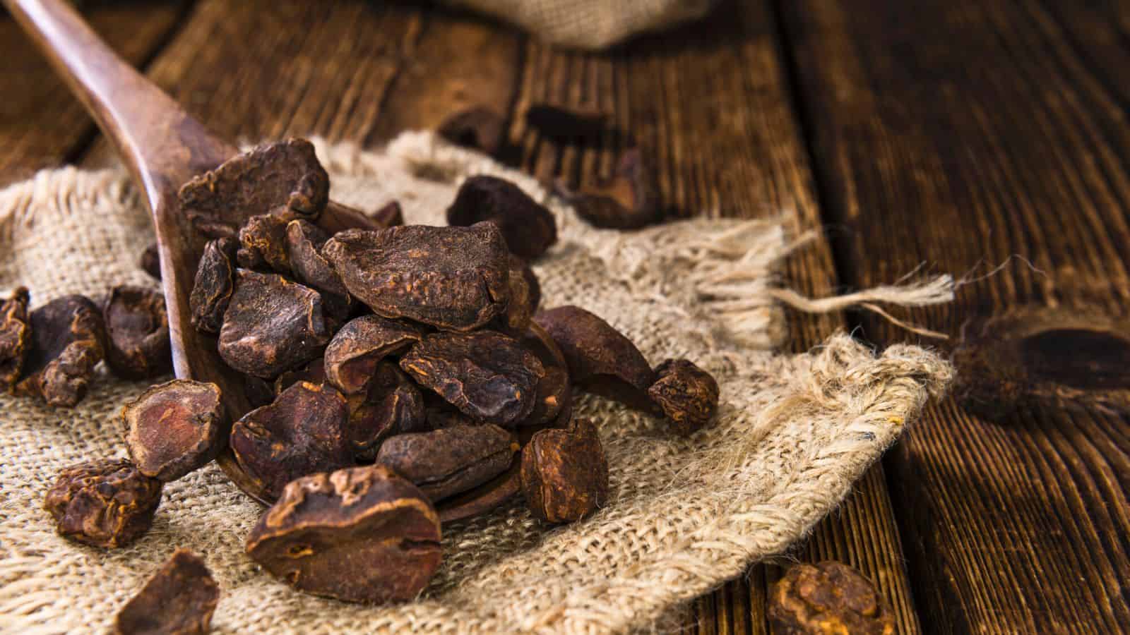 A wooden spoon holds several chunks of dried carob, resting on a piece of burlap fabric. The surface is a wooden table, and additional carob pieces are scattered around. The setting has a rustic feel.