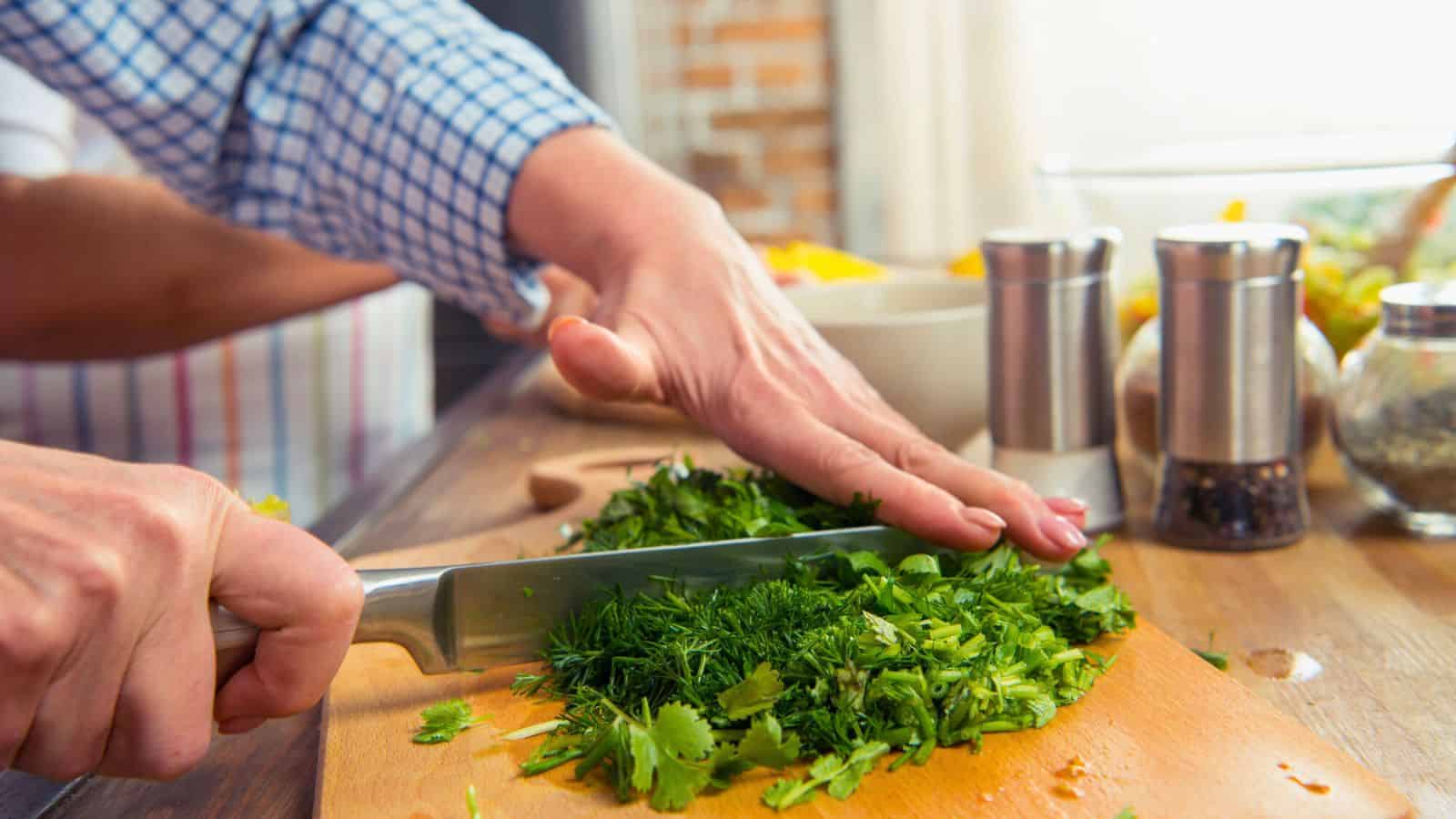 A person chops a variety of fresh green herbs on a wooden cutting board. A knife is in use, and nearby on the table are salt and pepper shakers, a jar, and a bowl. The person wears a checkered shirt and an apron.