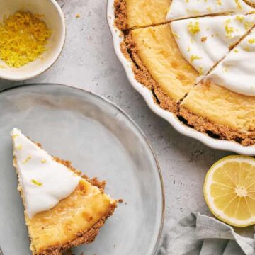 A lemon pie in a round dish with a few slices missing is on a gray surface. A slice is on a plate beside it, accompanied by a spoon. A lemon half, a bowl of zest, and a gray cloth are nearby.