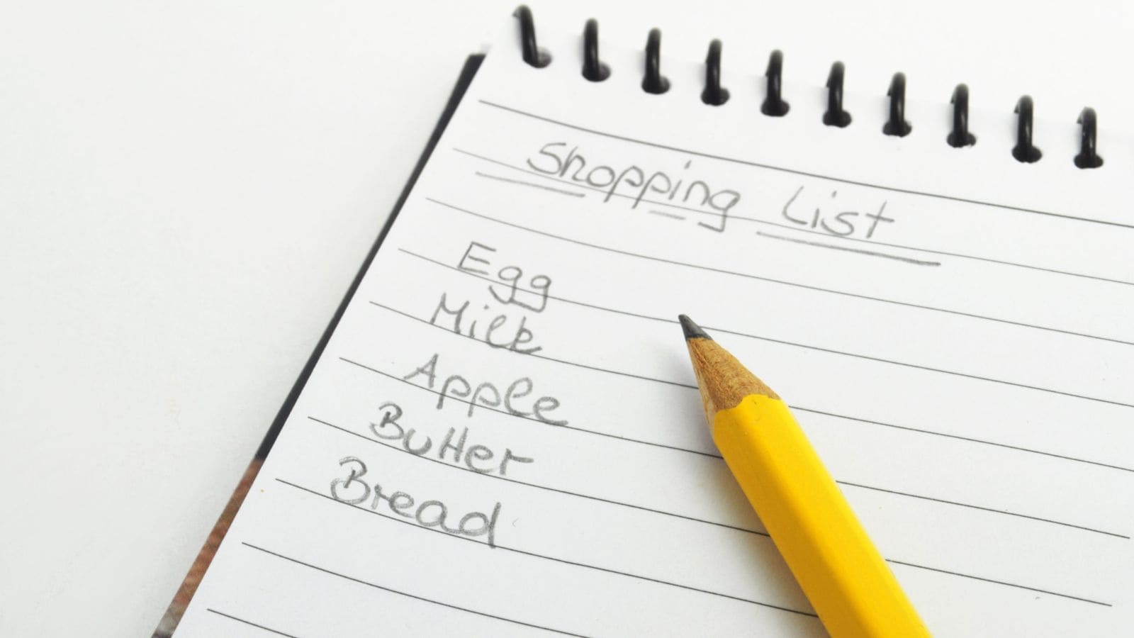 A spiral-bound notepad shows a handwritten shopping list. Items include egg, milk, apple, butter, and bread. A yellow pencil rests on the page, pointed towards the list.