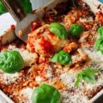 Close-up of a baked lasagna in a square dish, topped with grated cheese and fresh basil leaves. A serving spatula lifts a portion, revealing layers of pasta, sauce, and creamy cottage cheese. This delightful recipe is garnished with bright green basil.