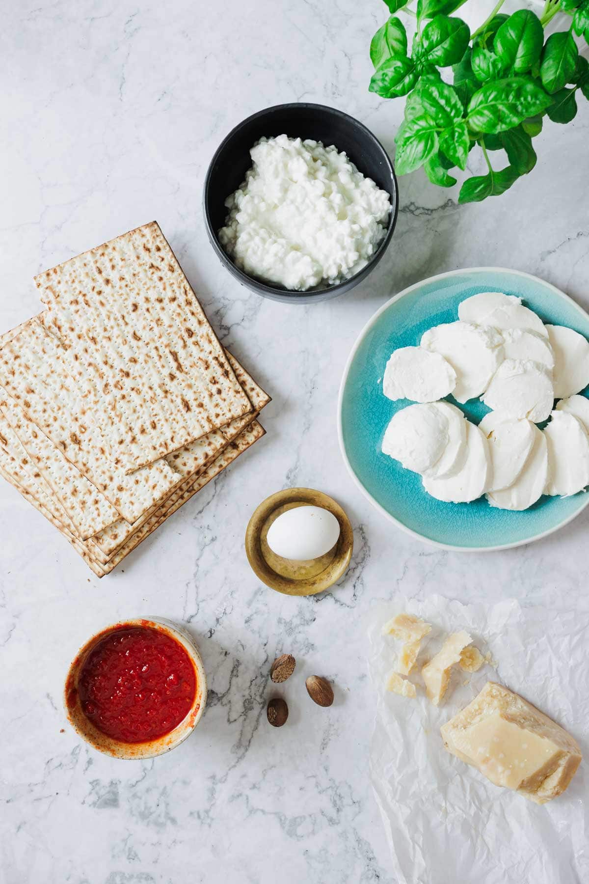 A marble surface showcases a Matzo Lasagna recipe with matzo crackers, a small bowl of cottage cheese, a blue plate with sliced mozzarella, red sauce in a bowl, an egg on a dish, whole nutmegs, parmesan, and fresh basil.