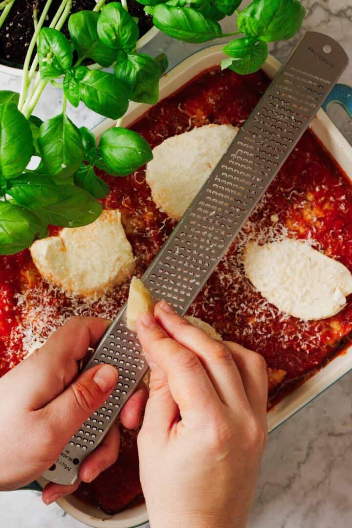 Close-up of hands grating cheese over a dish of baked pasta, reminiscent of a classic lasagna recipe, topped with tomato sauce and sliced cheese. Fresh basil plants are nearby, set elegantly on a marble countertop.
