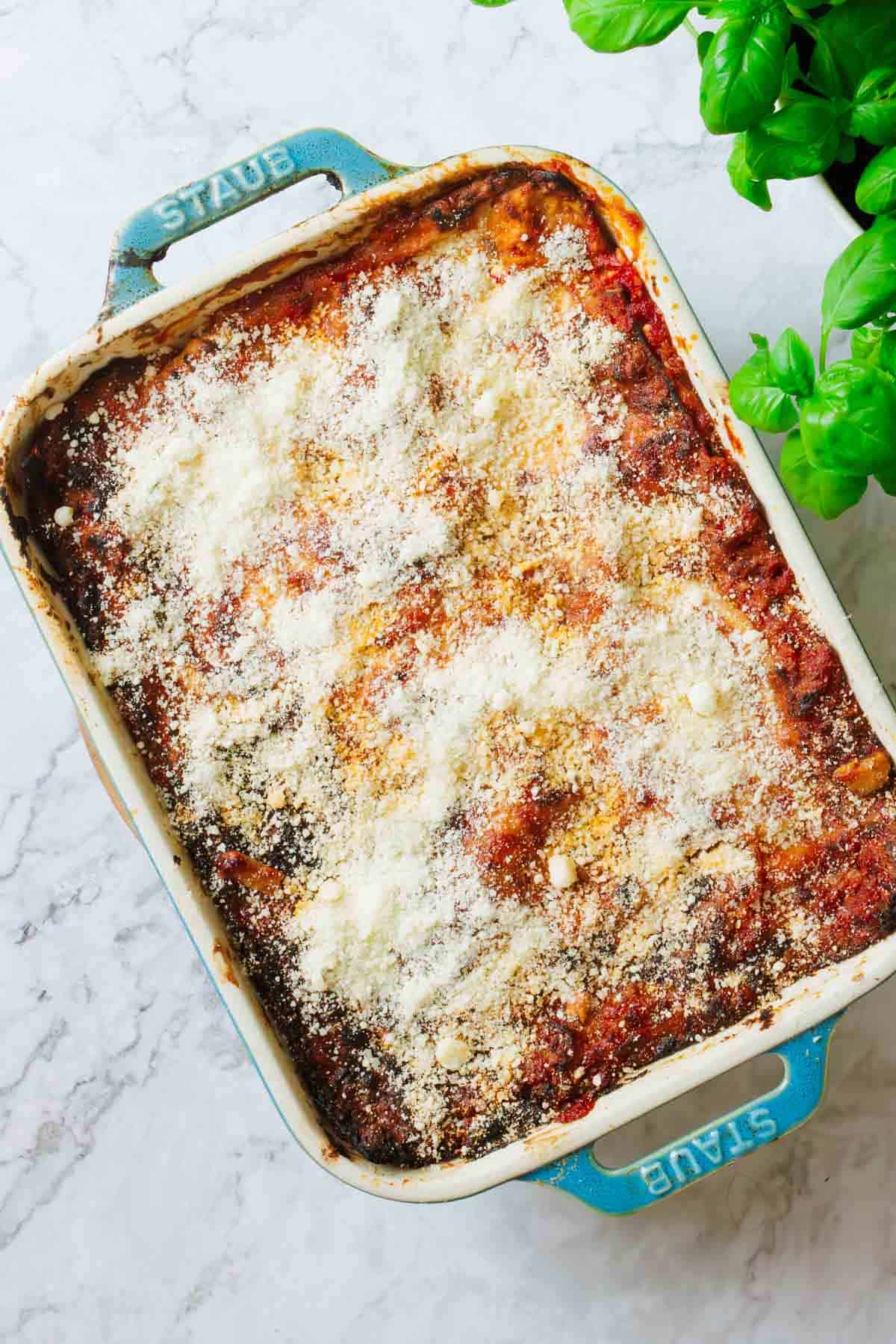 A rectangular baking dish filled with matzo lasagna topped with grated cheese sits on a marble surface. Fresh basil leaves are visible in the top right corner. The dish handles have the brand name "STAUB" on them, hinting at the unique twist of cottage cheese within this delightful creation.