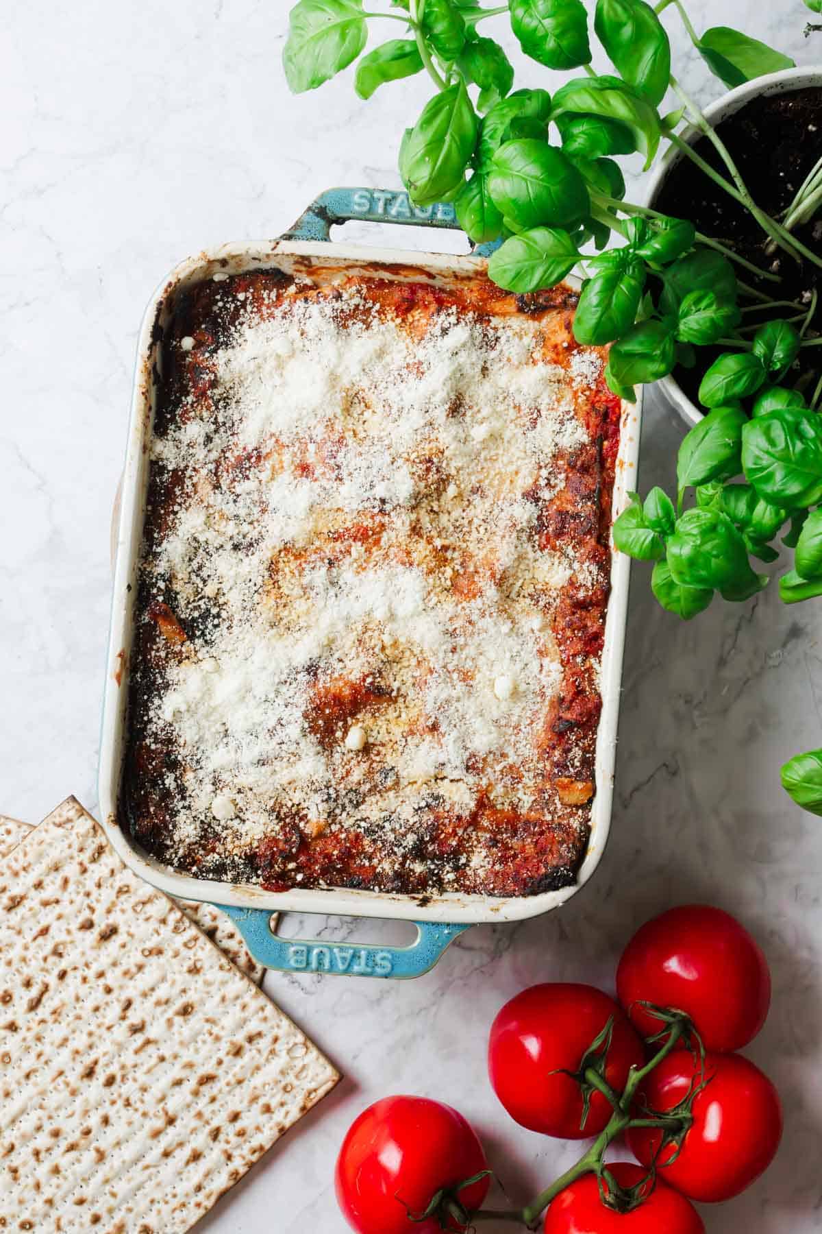 A baked matzo lasagna with a golden crust sprinkled with cheese in a rectangular blue dish. Beside it, fresh basil in a pot, three ripe tomatoes, and two pieces of matzo on a marble surface, creating the perfect scene for this delicious recipe.