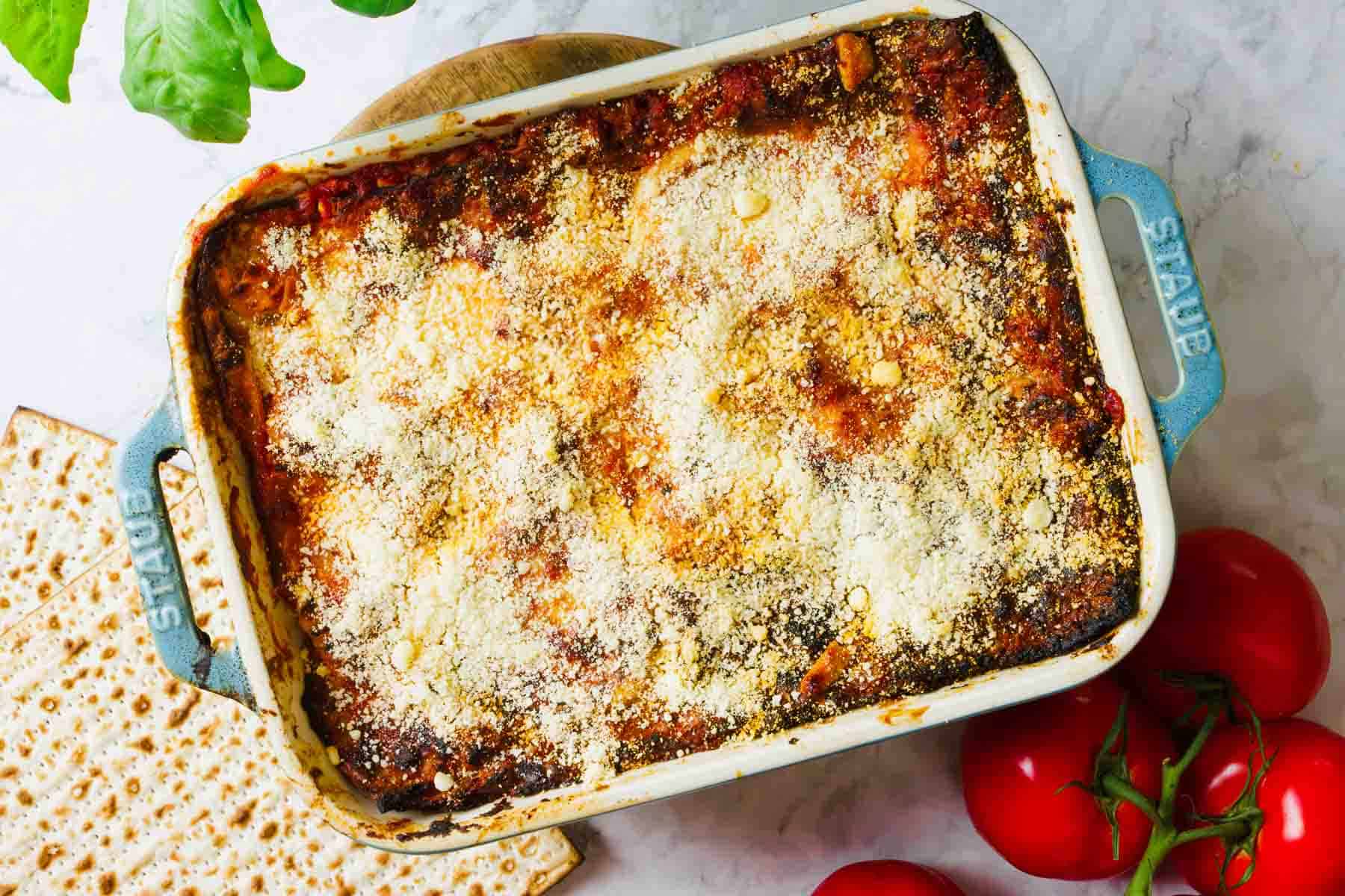 A baked casserole dish of matzo lasagna topped with golden-brown cheese and breadcrumbs. The dish sits on a marble surface, surrounded by fresh tomatoes, cottage cheese, and basil leaves.