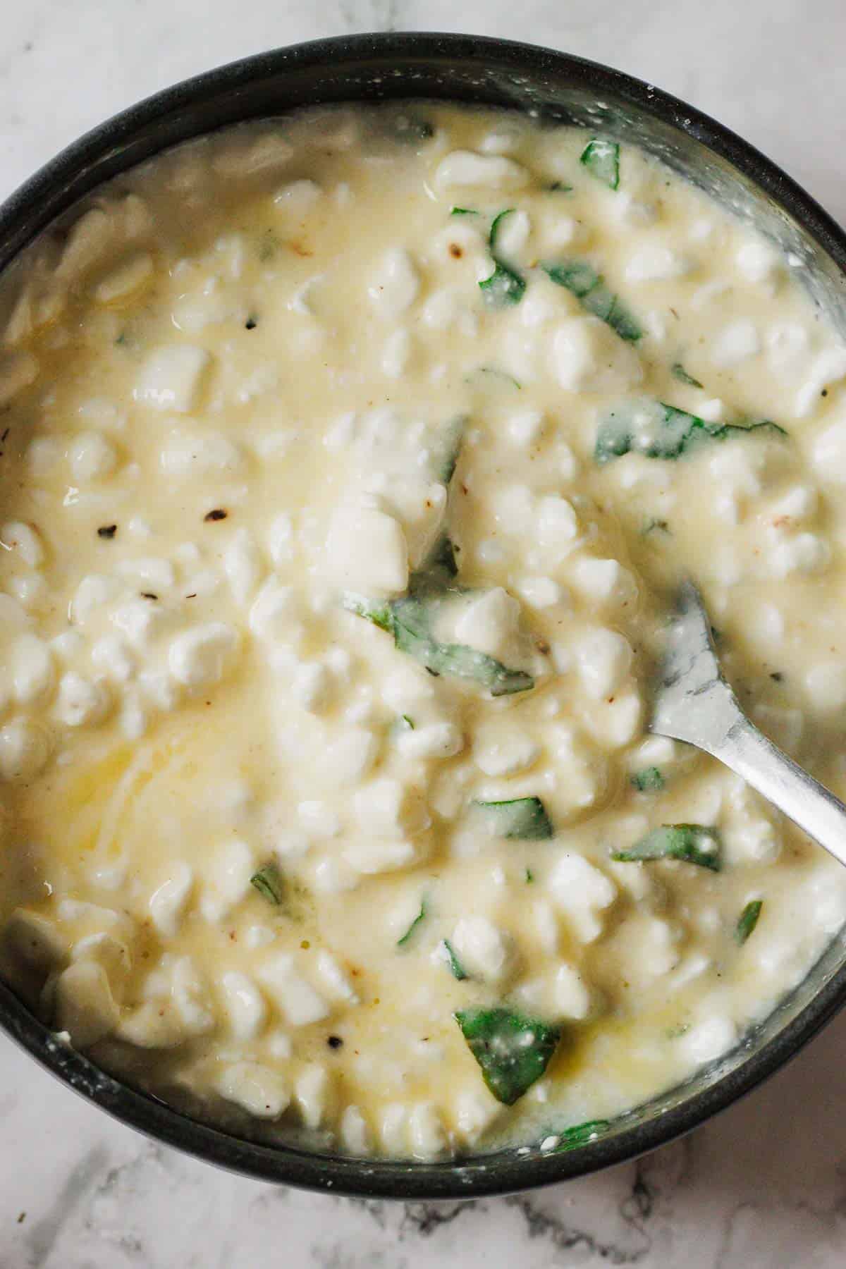 A bowl of creamy corn chowder with visible corn kernels and pieces of fresh green herbs. A spoon is partially submerged in the soup, which has a rich, thick texture reminiscent of a comforting matzo lasagna recipe. The bowl is set on a light-colored surface.