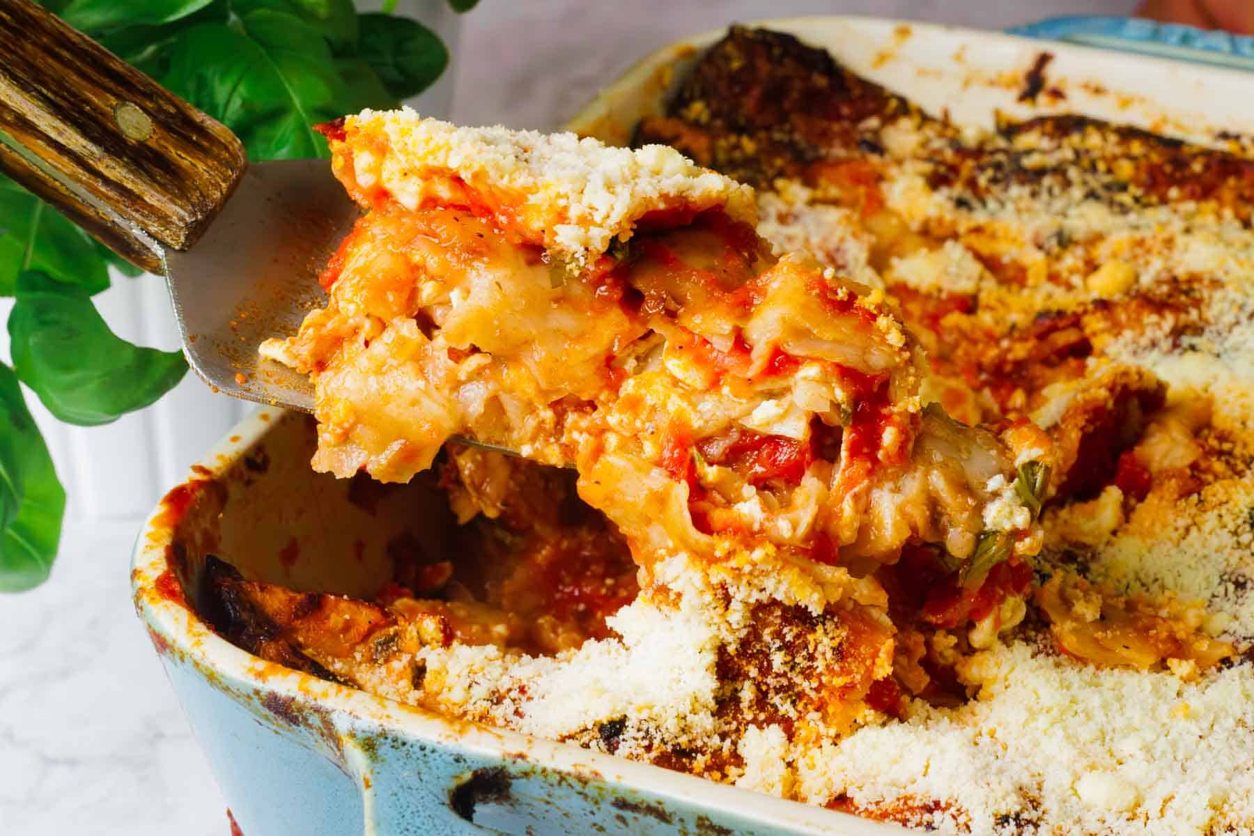 Close-up of a slice of Matzo Lasagna being lifted from a casserole dish with a spatula. This unique version is layered with cottage cheese, tomato sauce, and matzo sheets, topped with grated cheese. The dish has a golden-brown, crispy surface, while fresh basil adorns the backdrop.