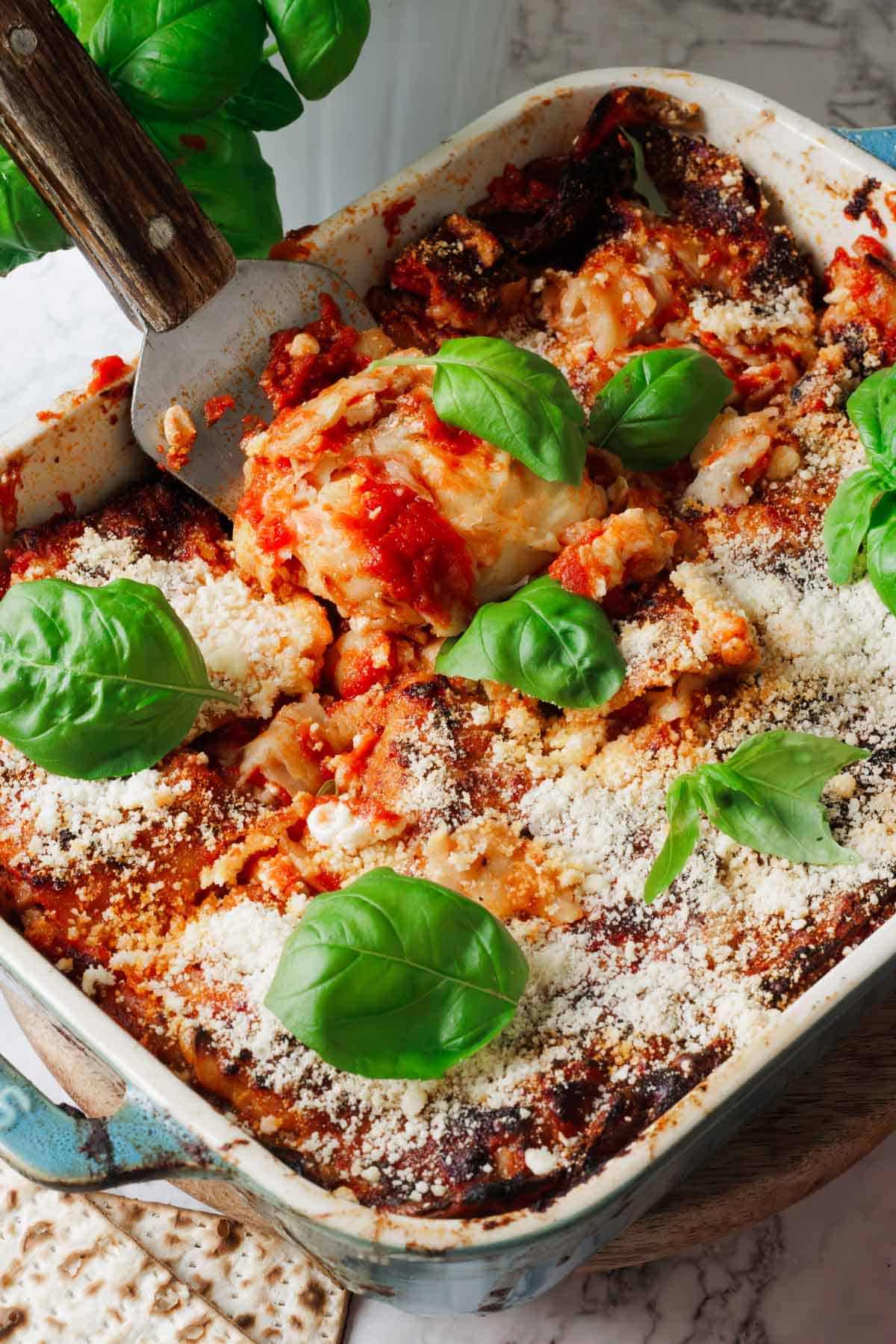 A baked dish of eggplant parmesan topped with melted cheese and marinara sauce, garnished with fresh basil leaves. A serving spatula is placed in the dish, lifting a portion. This delightful recipe sits on a light-colored surface, ready to enjoy.