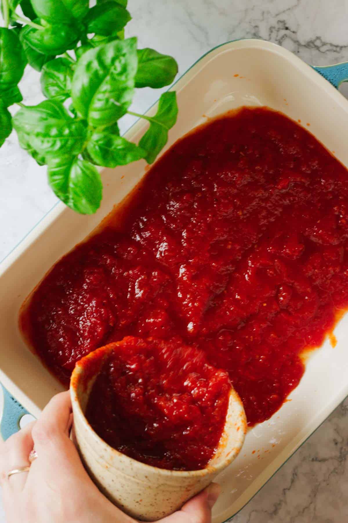 A beige baking dish filled with red tomato sauce sits ready for a delightful Matzo Lasagna. A hand pours more sauce from a small container, while a basil plant with green leaves watches from the top left corner over a light-colored marble surface.