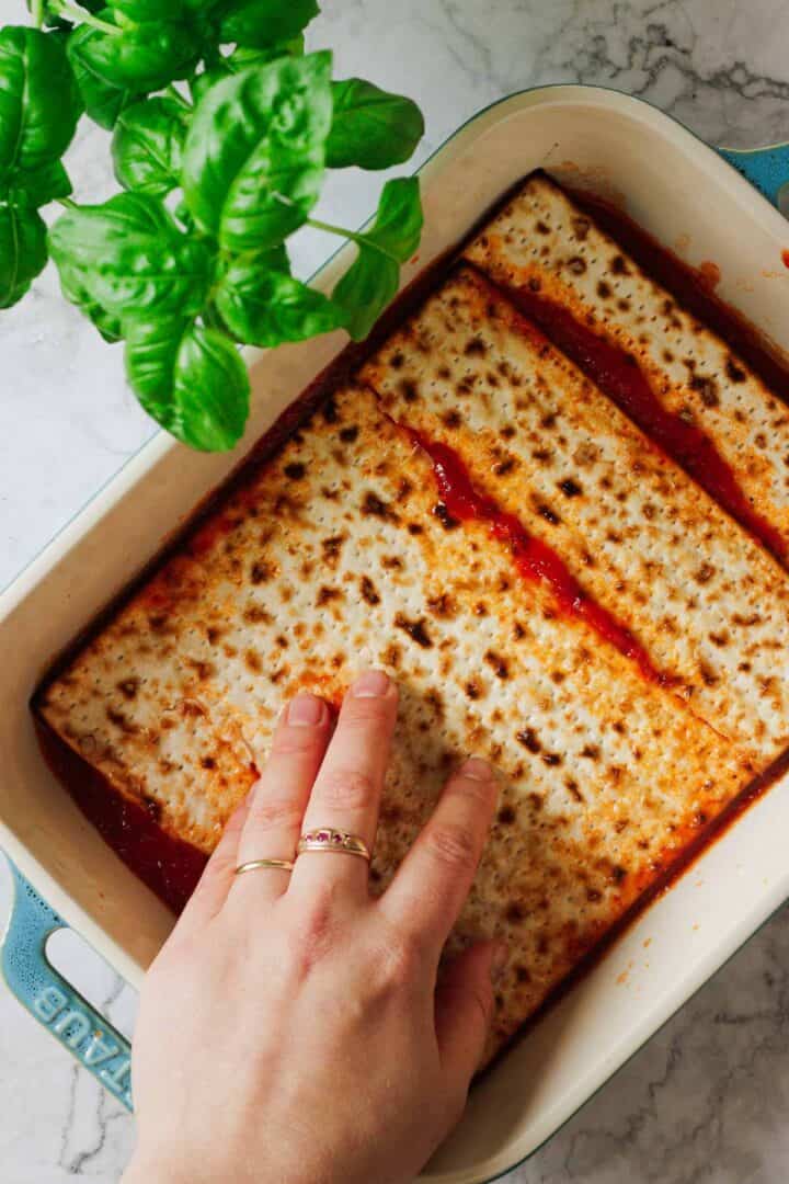A hand layers matzo sheets over tomato sauce in a blue rectangular baking dish, crafting a delightful Matzo Lasagna. Fresh green basil leaves and creamy cottage cheese are visible at the top, adding richness to this unique recipe on a marble countertop.