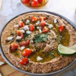 A round dish filled with creamy hummus, garnished with chopped tomatoes, onions, and herbs. A slice of lime is on the side. Olive oil is drizzled on top, and cherry tomatoes are visible in the background for a Mexican-style twist.