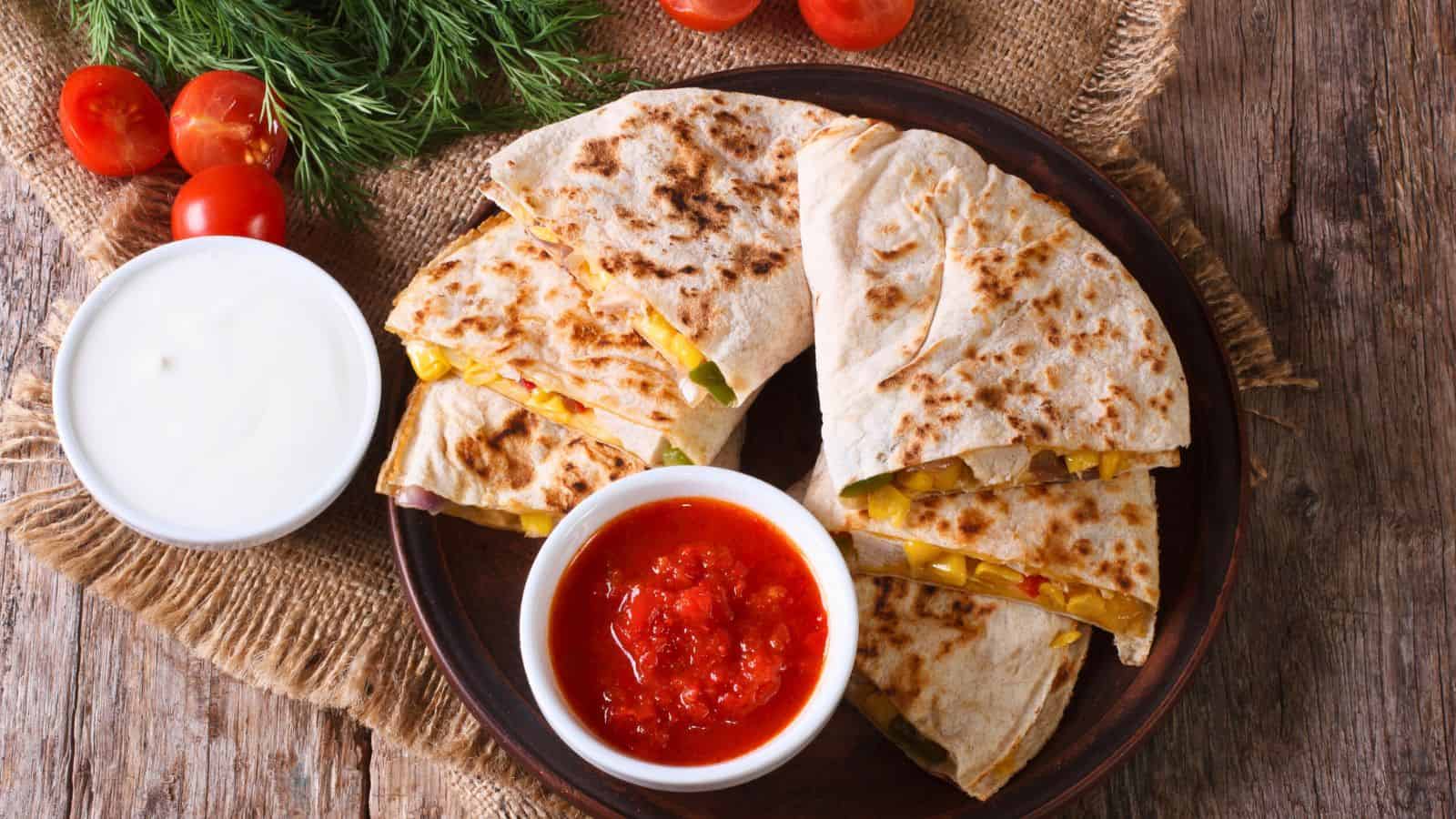 A wooden table displays a plate of sliced quesadillas with visible fillings like peppers and corn. A small bowl of red salsa and another of sour cream accompany the dish. Cherry tomatoes and dill lie nearby on a burlap cloth.