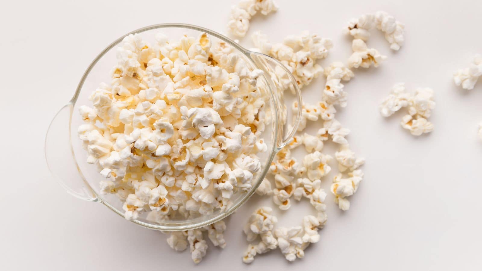 A glass bowl filled with popped popcorn sits on a white surface. Additional popcorn pieces are scattered around the bowl. The popcorn is fluffy and white, with some pieces showing a hint of golden brown.