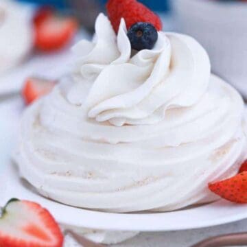 A homemade dessert delicately sits on a white plate—meringue topped with whipped cream, a raspberry, and a blueberry. Sliced and whole strawberries are artfully scattered around. Another plate and cup peek from the background, making it taste like mom's delightful touch.