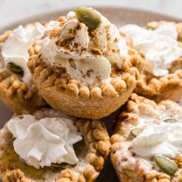 A plate with five small pies topped with whipped cream. The pies have decorative crimped edges, and some are sprinkled with seeds and cinnamon. The plate is set on a light-colored surface.