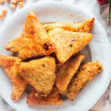 A white plate with several triangular fried pastries on a marble surface. Surrounding the plate are chickpeas, a red pepper, and cilantro leaves.