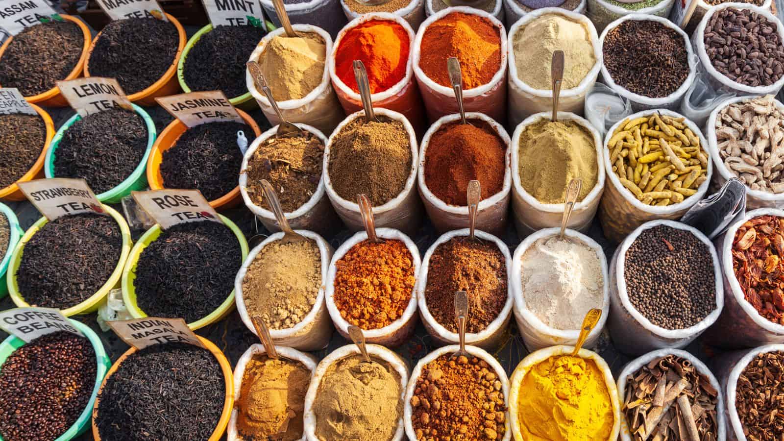 An array of open bags filled with various spices and teas, including turmeric, red chili, and Indian masala, are displayed at a market. Each bag has a metal scoop and is labeled. The spices are vibrant in color, while the teas are labeled in small signs.