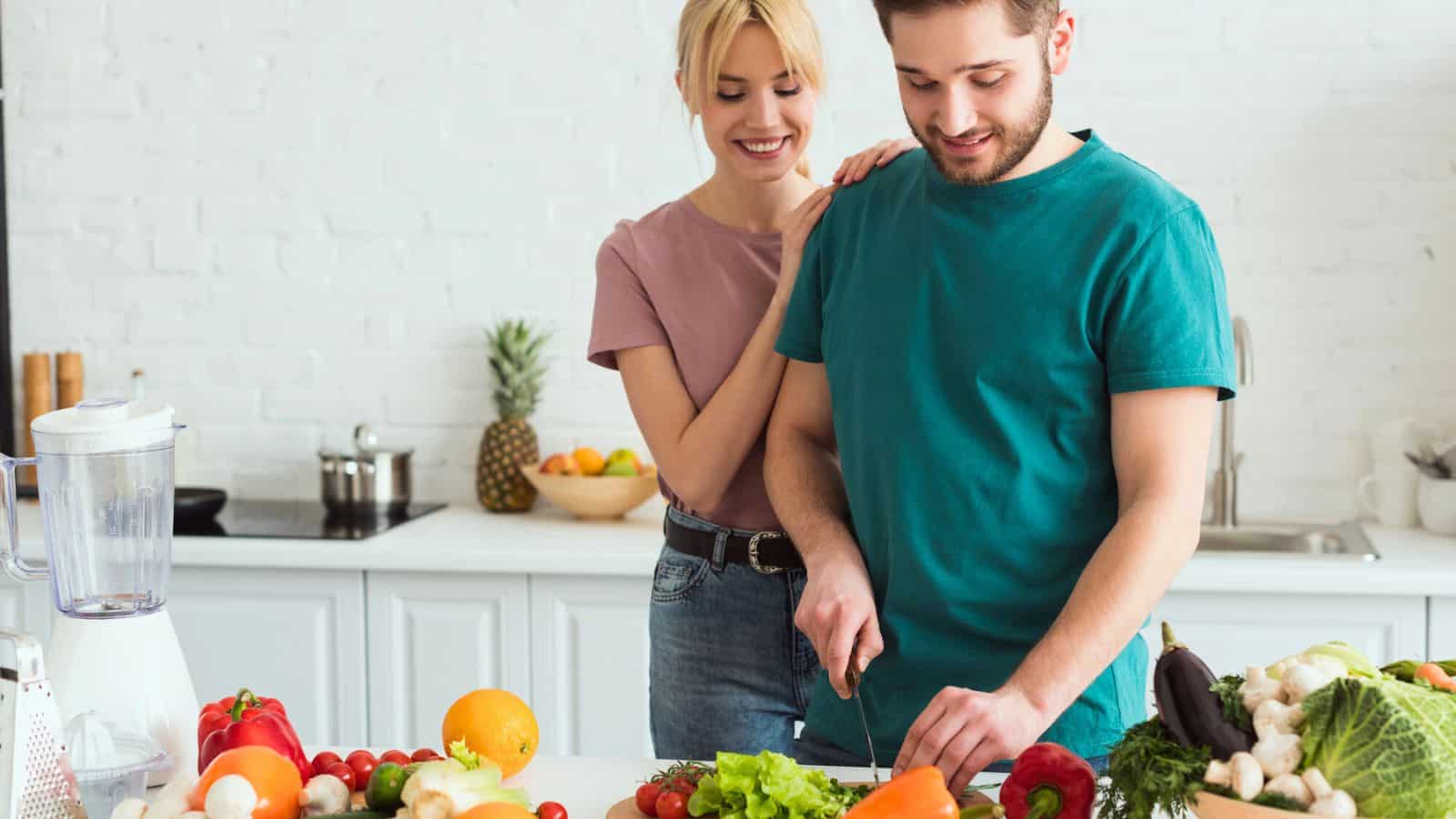 A man in a teal shirt slices a pepper on a kitchen counter filled with various fruits and vegetables. A woman in a pink shirt stands beside him, smiling. The kitchen features a blender and a modern white brick background.