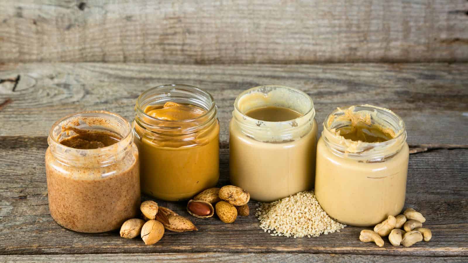 Four glass jars are filled with different nut and seed butters, placed on a rustic wooden surface. Whole nuts and seeds, including peanuts, sesame seeds, and cashews, are scattered around the jars.