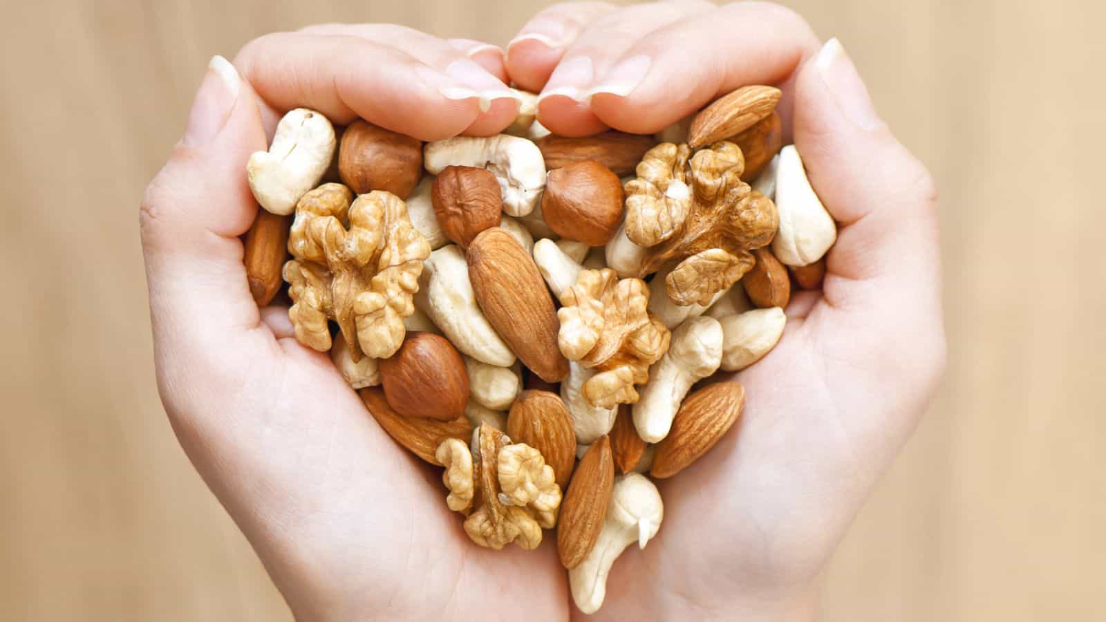 Two hands cupped together hold a variety of mixed nuts, including almonds, walnuts, cashews, and hazelnuts. The background is a neutral, blurred surface, emphasizing the assortment of nuts.