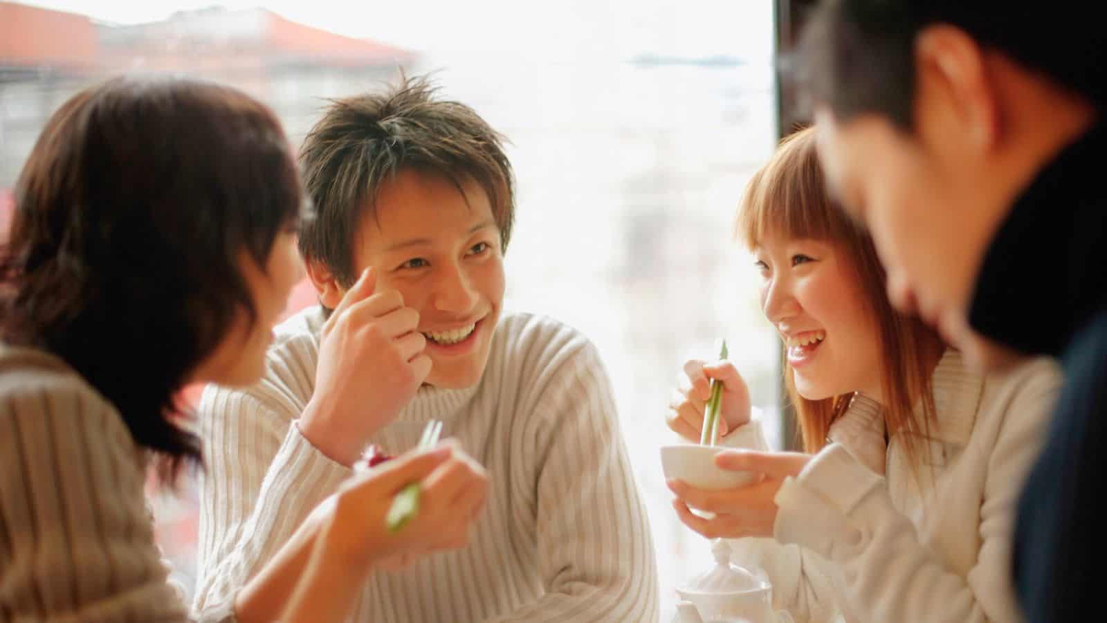 Four people sit together at a table, smiling and talking. Two hold cups, and the group appears relaxed and engaged in conversation. They are dressed in light-colored clothing. The background is softly blurred.