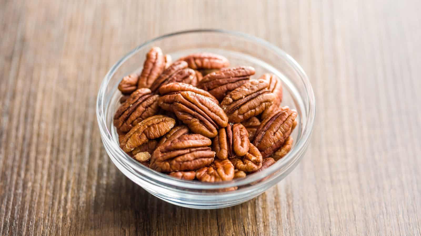 A glass bowl filled with shelled pecans sits on a wooden surface. The pecans have a reddish-brown color and are arranged neatly within the bowl. The wood grain of the surface is visible.