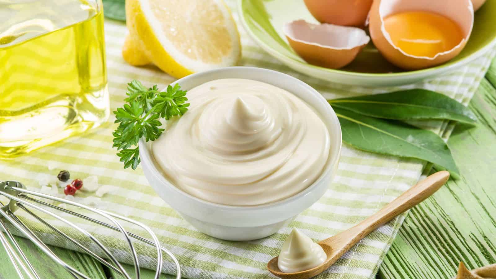 A bowl of creamy mayonnaise is garnished with parsley. Nearby are eggshells with yolk, a half lemon, a small wooden spoon, and a bottle of oil. A whisk and peppercorns rest on a green and white checkered cloth.