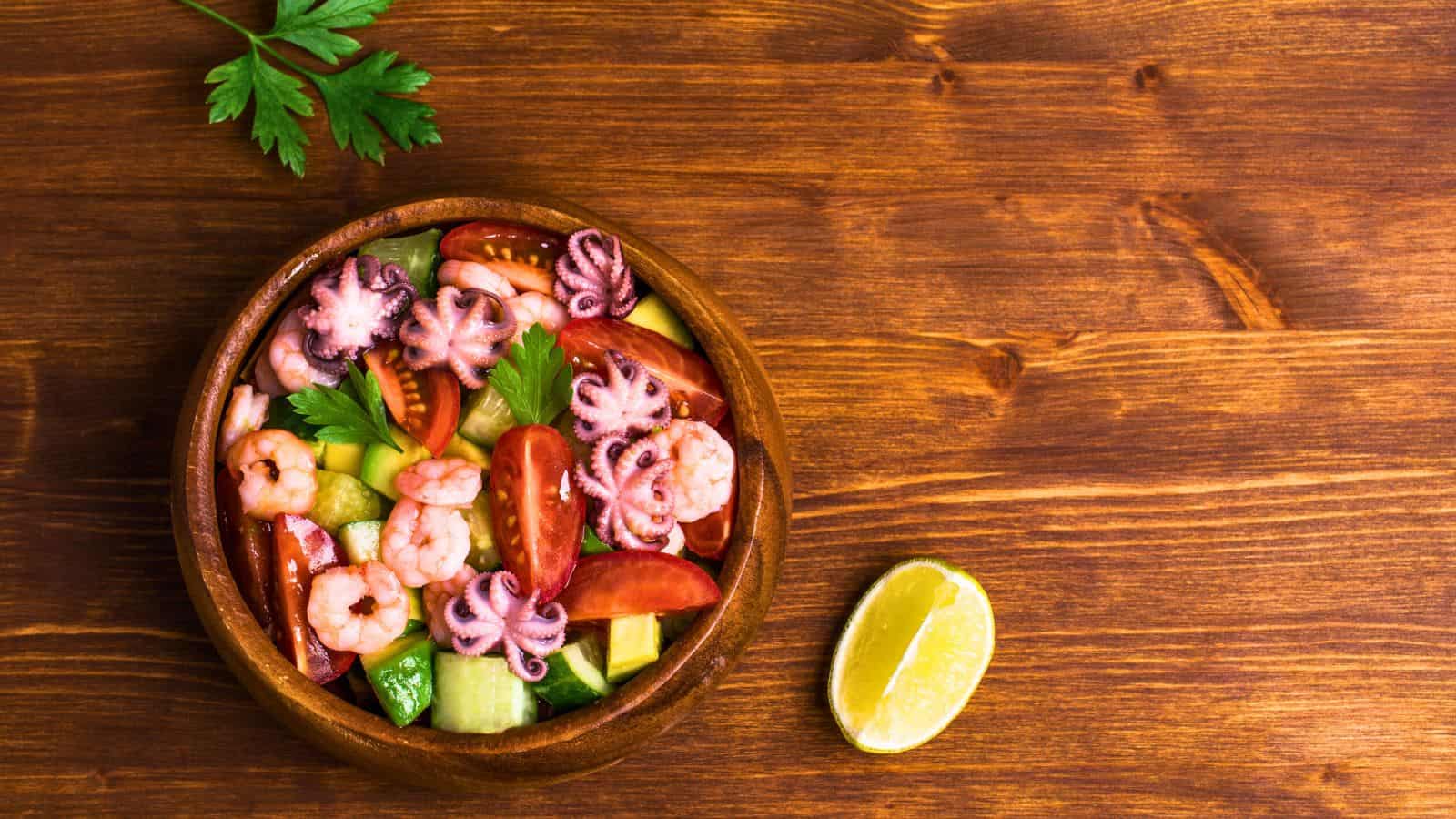 A wooden bowl containing a seafood salad is placed on a wooden surface. The salad includes shrimp, baby octopus, sliced tomatoes, cucumber, and parsley. A lime wedge is positioned to the right of the bowl.