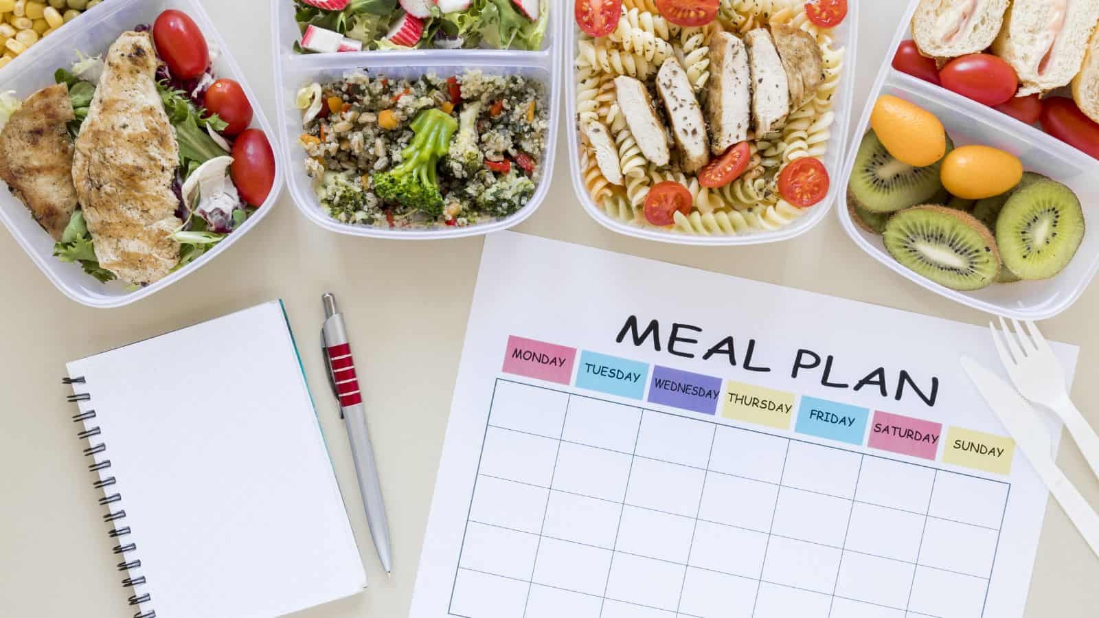 Top view of a meal prep setup. Clear containers with salads, grilled chicken, pasta, quinoa, and fruit slices are arranged around a blank meal plan chart. A spiral notebook and pen are also visible.