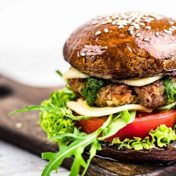 Indulge in this gourmet payday meal featuring a portobello mushroom bun, fresh lettuce, juicy tomato, a beef patty, and cheese with green sauce. Topped with sesame seeds, this delicious creation rests invitingly on a wooden board against a light backdrop.