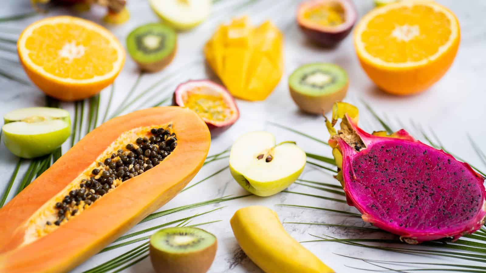A variety of sliced fruits are arranged on a white surface. Visible are papaya, dragon fruit, oranges, apples, starfruit, bananas, kiwis, and passion fruits. Palm leaves are scattered among the fruits as decoration.